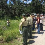 2018 annual workshop A group of people, some in state agency uniforms, watch a woman present about aquatic plants while standing in a pond.