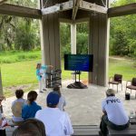 2022 workshop_Nancy Bissett A group of people watch a presentation by a woman while sitting in an outdoor amphitheater.