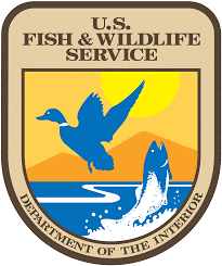 US Fish and Wildlife Service logo featuring a blue silhouette of a duck, fish, and lake, with orange mountains and a yellow sun in the background encircled by a badge shaped tan and brown outline with the words US Fish and Wildlife Service above and "Department of the Interior" below.