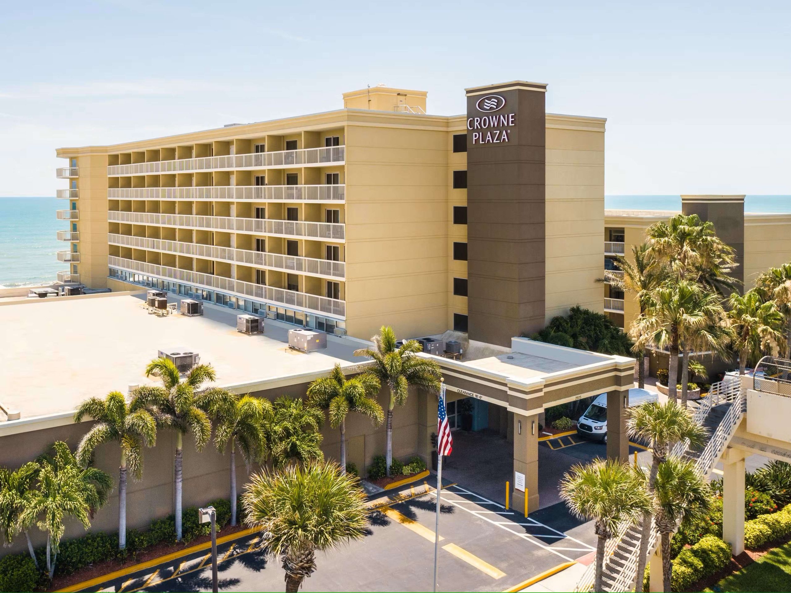 A yellow-brown six-story building with a row of balconies is in the foreground. This is the Crowne Plaza hotel in Melbourne, FL. There are palm trees around the building and the ocean in the background, showing that the building is on the beach.